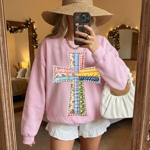 Op de afbeelding: Een lichtroze sweatshirt met een kleurrijke kruisapplicatie. Het kruis is gemaakt van verschillende stoffen met patronen, waaronder bloemen, ruitjes en stippen. De persoon draagt een strohoed, gouden sieraden en een witte short.