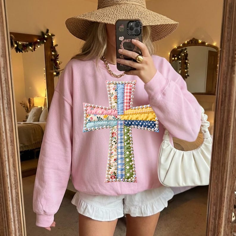 Op de afbeelding: Een lichtroze sweatshirt met een kleurrijke kruisapplicatie. Het kruis is gemaakt van verschillende stoffen met patronen, waaronder bloemen, ruitjes en stippen. De persoon draagt een strohoed, gouden sieraden en een witte short.