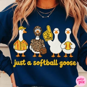 May include: Navy blue sweatshirt with a playful design featuring four cartoon geese dressed in softball gear. One goose wears a baseball uniform, another a leopard-print outfit, and the third holds a foam finger. The text "just a softball goose" is printed in yellow.
