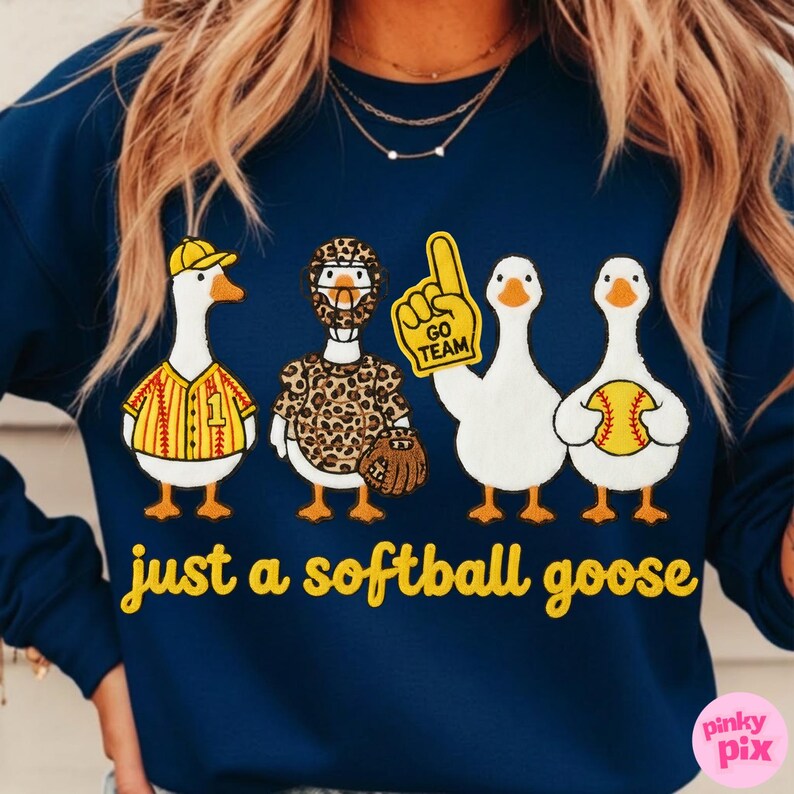May include: Navy blue sweatshirt with a playful design featuring four cartoon geese dressed in softball gear. One goose wears a baseball uniform, another a leopard-print outfit, and the third holds a foam finger. The text "just a softball goose" is printed in yellow.