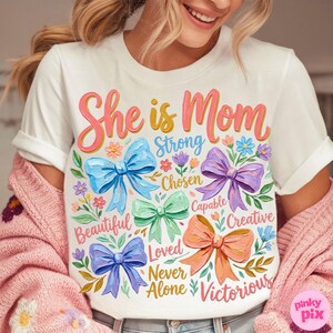 K&ouml;nnte beinhalten: Wei&szlig;es T-Shirt mit dem Text "She is Mom" in rosa und gelber Schrift. Das Shirt zeigt bunte Schleifen und florale Akzente mit W&ouml;rtern wie "Strong", "Beautiful" und "Victorious".
