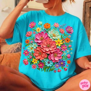 May include: A turquoise t-shirt featuring a vibrant floral design. The bouquet includes pink, orange, yellow, and purple flowers with green leaves. The shirt has short sleeves and a round neckline. The image also includes a pink and white logo in the bottom right corner.