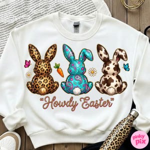 May include: White sweatshirt featuring three Easter bunnies with unique patterns: leopard print, teal paisley, and brown and white spots. The phrase "Howdy Easter" is written in a decorative font below the bunnies. Butterflies, carrots, and daisies are also included.