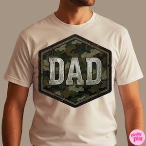 Pode incluir: Camiseta branca com um design de camuflagem e a palavra "DAD" em uma fonte met&aacute;lica desgastada. O gr&aacute;fico est&aacute; dentro de uma moldura hexagonal. Uma pe&ccedil;a de roupa casual.