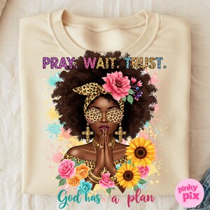 May include: A beige t-shirt with a colorful illustration of a woman with an afro, wearing sunglasses and a leopard print top. The text "Pray Wait Trust. God has a plan" is printed on the shirt.