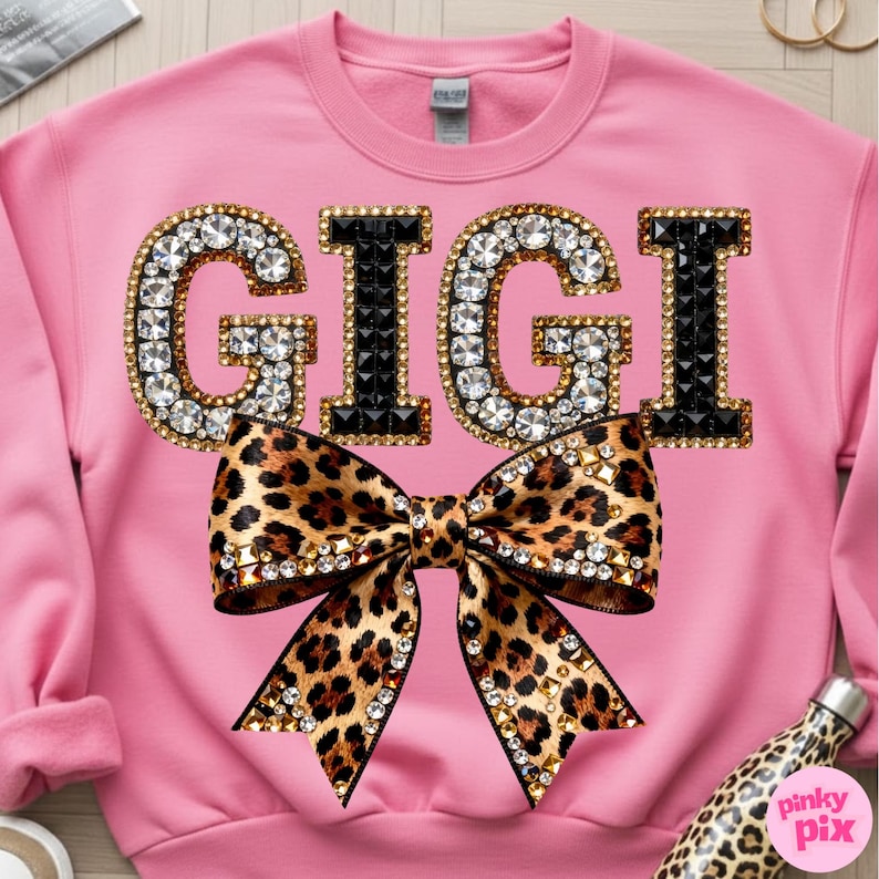 May include: Pink sweatshirt with the name "GIGI" in jeweled letters and a leopard print bow. The letters are outlined in gold and encrusted with clear rhinestones and black gems. The bow is also adorned with rhinestones.