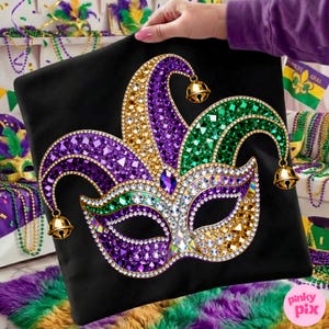 May include: Black pillow featuring a jeweled Mardi Gras mask design. The mask is adorned with purple, green, gold, and clear crystals, and includes small gold bells. The pillow is held by a person.