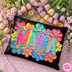 May include: Black decorative pillow with the word "MAMA" in colorful, textured lettering, surrounded by vibrant, painted flowers in shades of pink, yellow, blue, and orange. The pillow is set against a background of flowers.