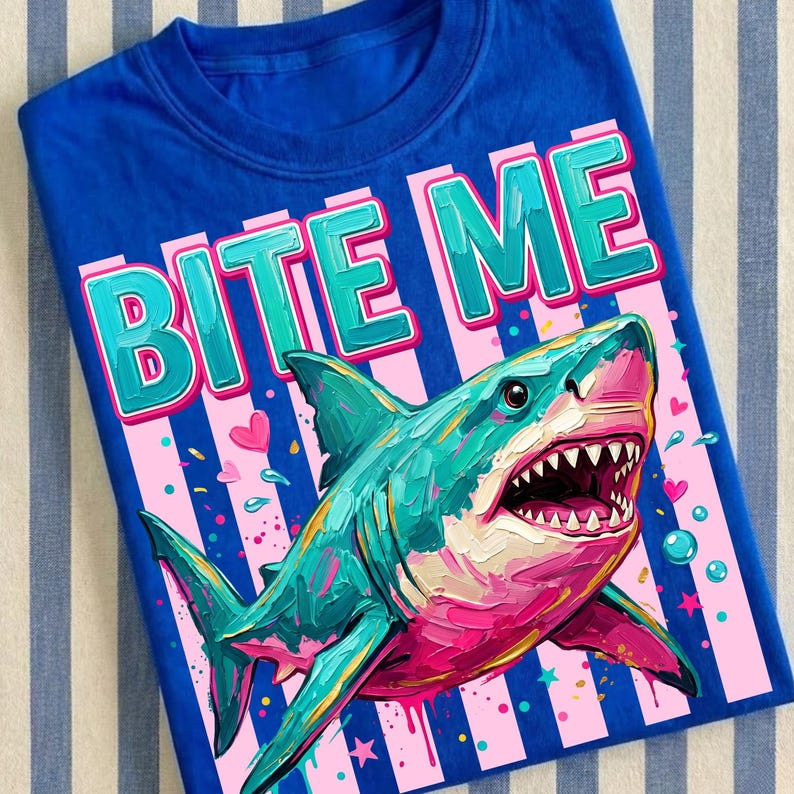 May include: A blue t-shirt featuring a colorful shark illustration with the words "BITE ME" in a playful font. The shark is painted in shades of teal, pink, and white, with visible teeth. The design includes pink and white stripes and hearts.