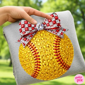 May include: A softball adorned with yellow rhinestones and red stitching, topped with a bow featuring red and white checkered accents and a central diamond. The softball is resting on a grey surface.