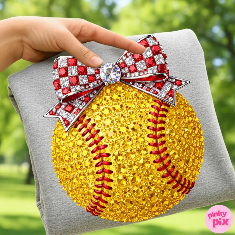 May include: A softball adorned with yellow rhinestones and red stitching, topped with a bow featuring red and white checkered accents and a central diamond. The softball is resting on a grey surface.