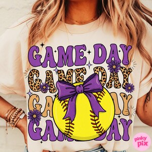 May include: Cream-colored t-shirt with a softball graphic. The design features the words "GAME DAY" in purple and leopard print, with a yellow softball and purple bow. The shirt has a casual, sporty theme.