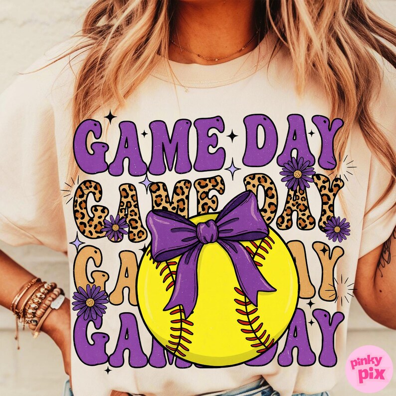 May include: Cream-colored t-shirt with a softball graphic. The design features the words "GAME DAY" in purple and leopard print, with a yellow softball and purple bow. The shirt has a casual, sporty theme.
