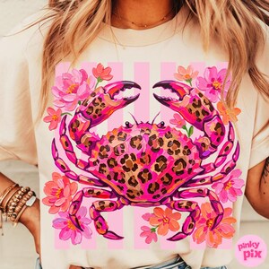 May include: A light-colored t-shirt featuring a vibrant pink crab with a leopard print design. The crab is surrounded by pink and orange flowers against a background of pink stripes. The shirt has a relaxed fit.