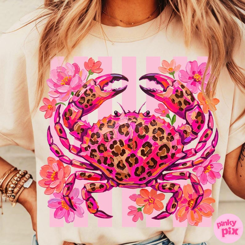 May include: A light-colored t-shirt featuring a vibrant pink crab with a leopard print design. The crab is surrounded by pink and orange flowers against a background of pink stripes. The shirt has a relaxed fit.