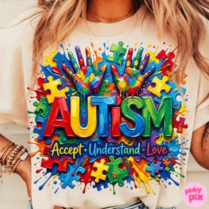 Op de afbeelding: Cr&egrave;mekleurig T-shirt met een kleurrijk ontwerp. Het woord "AUTISM" staat in grote, 3D-letters afgebeeld, omringd door puzzelstukjes, handafdrukken en de woorden "Accept, Understand, Love". Het ontwerp is omgeven door verfspatten.