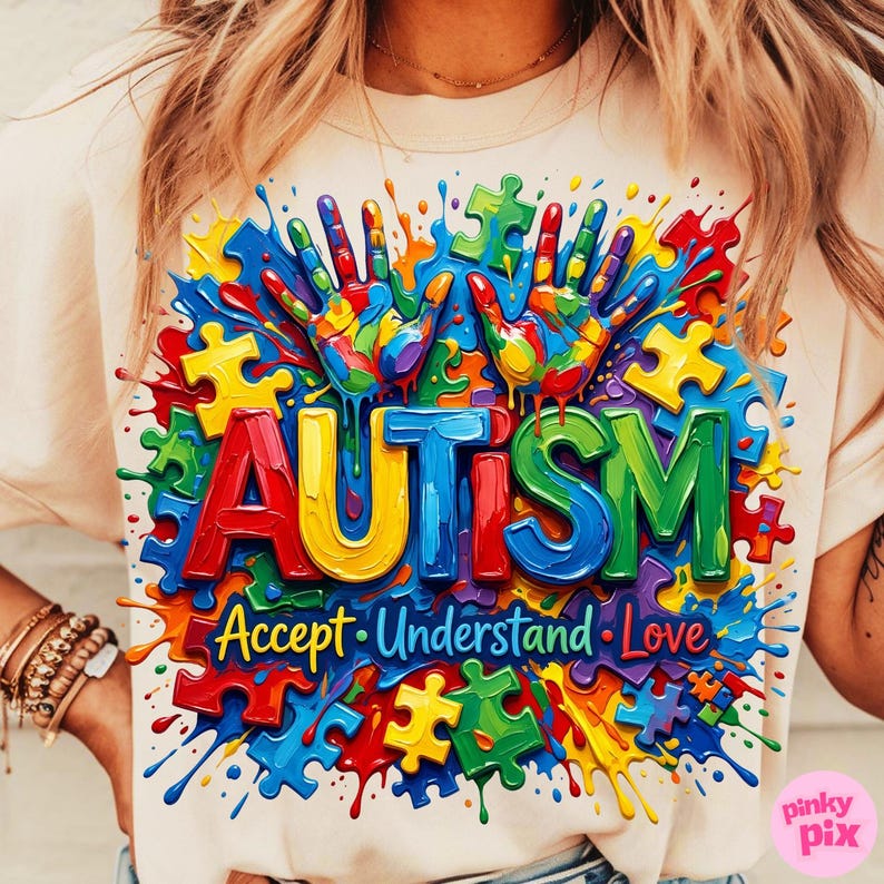Op de afbeelding: Cr&egrave;mekleurig T-shirt met een kleurrijk ontwerp. Het woord "AUTISM" staat in grote, 3D-letters afgebeeld, omringd door puzzelstukjes, handafdrukken en de woorden "Accept, Understand, Love". Het ontwerp is omgeven door verfspatten.