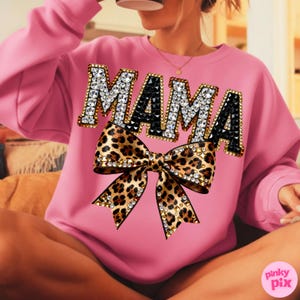 May include: Pink sweatshirt with the word "MAMA" in rhinestone-studded letters and a leopard print bow. The sweatshirt is a solid pink color and has a crew neck. The bow is embellished with rhinestones.