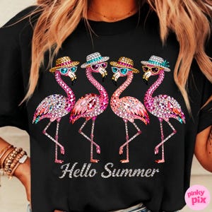 May include: Black t-shirt featuring four pink flamingo illustrations with sunglasses and hats. The flamingos are embellished with sparkling details. The text "Hello Summer" is displayed in a silver font.