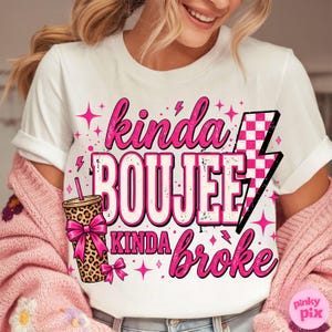 May include: A white short-sleeve t-shirt featuring a graphic design with the text "kinda BOUJEE kinda broke" in pink and black. The design includes a leopard print tumbler with a pink bow and a pink and white checkered lightning bolt icon.