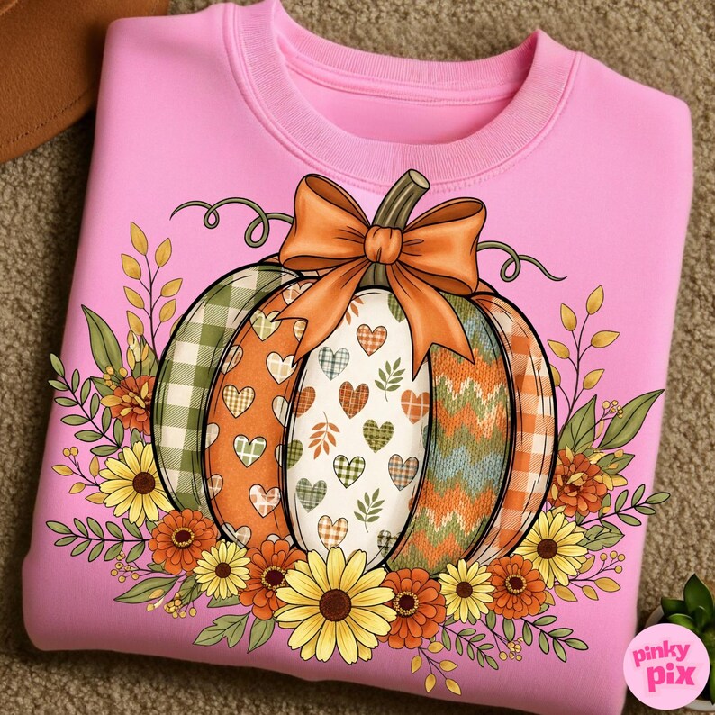 May include: Pink sweatshirt featuring a colorful pumpkin design. The pumpkin has sections with plaid, hearts, and patterned textures, topped with an orange bow. Floral accents and leaves surround the pumpkin, creating a fall-themed aesthetic.