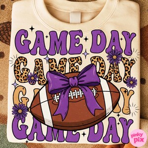 May include: Cream-colored sweatshirt with a retro design featuring the words "GAME DAY" in purple, leopard print, and tan. A brown football with a purple bow is in the center. The design includes purple flowers and stars.