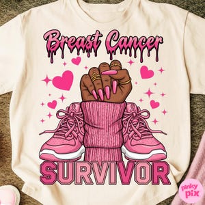 May include: Cream-colored t-shirt with a graphic design. The design features the text "Breast Cancer" in pink dripping letters above a clenched fist with pink nails and rings. Pink sneakers and the word "SURVIVOR" are below the fist.
