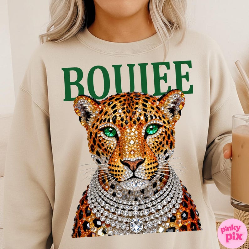 May include: Beige sweatshirt with a leopard graphic wearing a diamond necklace and green eyes. The word "BOUIEE" is printed in green above the leopard's head. The sweatshirt is a crew neck style.