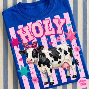 May include: A royal blue t-shirt featuring a cow illustration with a leopard print headband. The word "HOLY" is printed in pink above the cow. Pink stars, hearts, and flowers decorate the shirt.