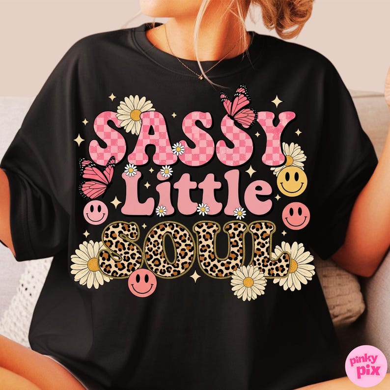 May include: Black t-shirt with the words "SASSY Little SOUL" in pink, leopard print, and white lettering. The design includes pink butterflies, daisies, and smiley faces. A fun, retro-style graphic tee.