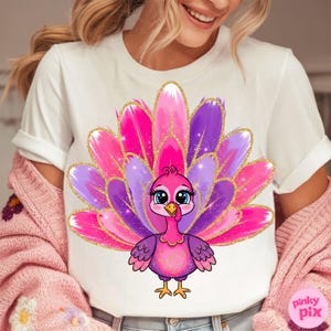 May include: White t-shirt featuring a cartoon turkey with a vibrant pink, purple, and gold feather design. The turkey has large blue eyes and is centered on the shirt. The text "pinky pix" is in the lower right corner.