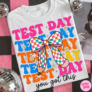 May include: White t-shirt with a colorful graphic design. The design features the words "TEST DAY" in pink, blue, and orange, along with a checkered bow and pencil illustration. The phrase "you got this" is also included.