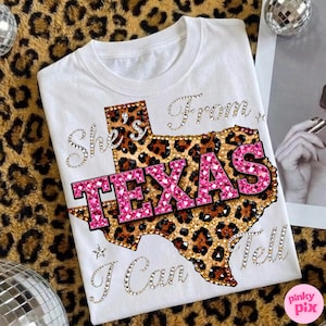 May include: White t-shirt with a leopard print Texas outline and the text "TEXAS" in pink rhinestones. The shirt also features the phrases "She's From" and "I Can Tell" in rhinestone lettering.