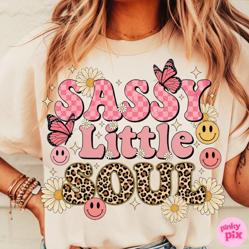 May include: Cream-colored t-shirt with the text "SASSY Little SOUL" in pink, checkered, and leopard print. The design includes butterflies, daisies, and smiley faces. A fun and colorful graphic tee.