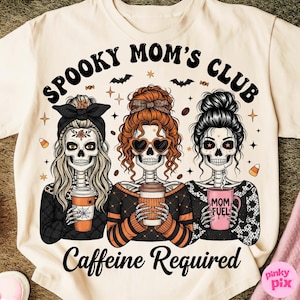 Puede incluir: Camiseta color crema con un diseño de Halloween. El diseño presenta a tres mujeres esqueleto sosteniendo tazas de café, con el texto "Spooky Mom's Club" y "Caffeine Required". Murciélagos y estrellas completan el diseño.