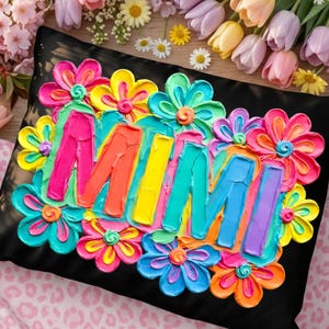 May include: Black decorative pillow with the name "MIMI" in colorful, textured paint, surrounded by vibrant, multi-colored flowers. The pillow is a square shape, and the flowers are in shades of pink, yellow, blue, and orange.