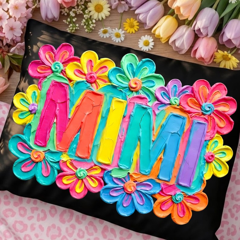 May include: Black decorative pillow with the name "MIMI" in colorful, textured paint, surrounded by vibrant, multi-colored flowers. The pillow is a square shape, and the flowers are in shades of pink, yellow, blue, and orange.