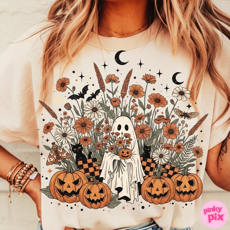 May include: Cream-coloured t-shirt with a Halloween graphic. The design features a ghost holding a pumpkin, surrounded by flowers, pumpkins, black cats, bats, and moons. The artwork has a vintage, botanical style.