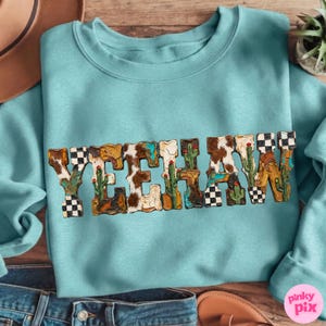 Może przedstawiać: Jasnoniebieska bluza z napisem "YEHAW" w ozdobnej czcionce. Litery zaprojektowano w stylu western, z wykorzystaniem skóry krowiej, kaktusów i kowbojskich butów. Bluza wykonana jest z miękkiego materiału.