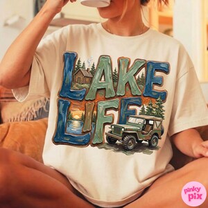May include: Cream-colored t-shirt with a colorful "LAKE LIFE" graphic. The design features a cabin, lake, sunset, and a green jeep. The text is in blue and green, with orange accents. The shirt is a casual wear item.