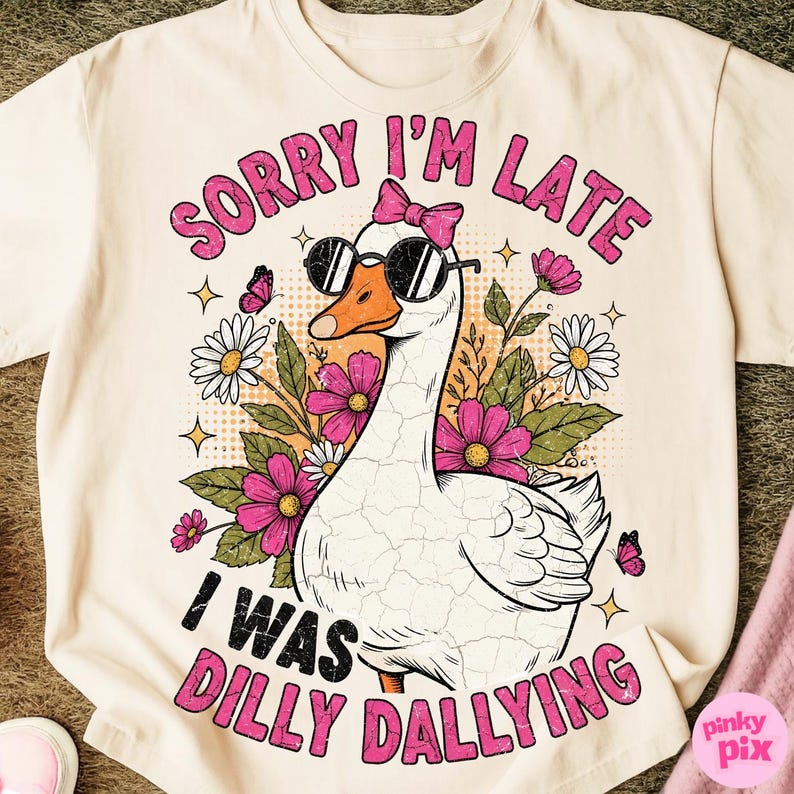 K&ouml;nnte beinhalten: Cremefarbenes T-Shirt mit einer Cartoon-Gans, die eine Sonnenbrille und eine rosa Schleife tr&auml;gt. Die Gans ist von Blumen und Schmetterlingen umgeben, mit dem Text "SORRY I'M LATE I WAS DILLY DALLYING" in Pink.