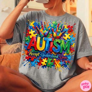 Op de afbeelding: Grijs t-shirt met een kleurrijk grafisch ontwerp. Het ontwerp toont het woord "AUTISME" in vetgedrukte, veelkleurige letters, omringd door puzzelstukjes en handafdrukken. De tekst "Accepteer. Begrijp. Liefde." staat eronder.