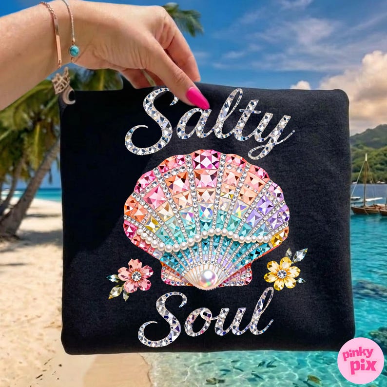 May include: Black tote bag with the words "Salty Soul" in a sparkling font. The bag features a colorful seashell design with sparkling accents and floral embellishments. The background shows a beach scene.