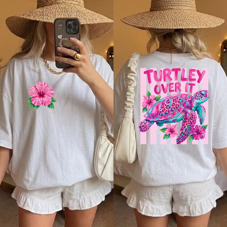 May include: White t-shirt with a pink hibiscus flower graphic on the front. The back of the shirt features a colorful turtle design with the text "Turtley Over It" in pink. The shirt is paired with white shorts.