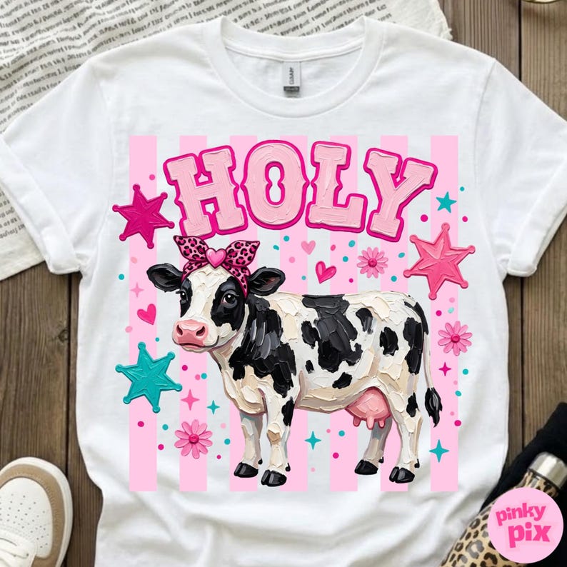 May include: White t-shirt featuring a cartoon cow with black and white spots, wearing a pink bow and a pink headband. The word "HOLY" is above the cow in pink letters. Pink stars, hearts, and flowers decorate the design.
