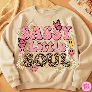 May include: Beige sweatshirt with the text "SASSY Little SOUL" in pink, checkered, and leopard print lettering. The design includes pink butterflies, daisies, and smiley faces. A casual, fun, and retro-inspired design.