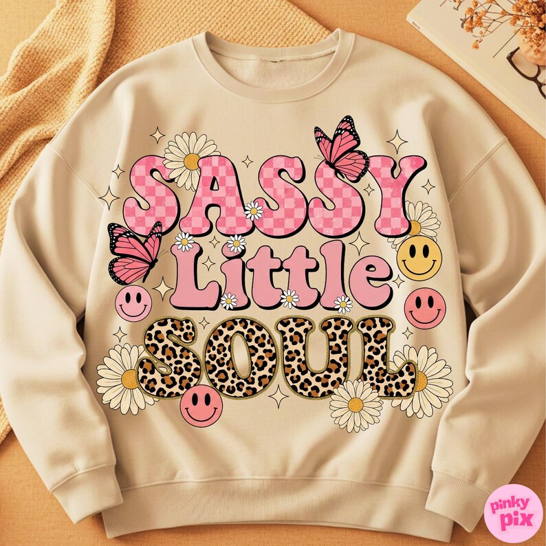 May include: Beige sweatshirt with the text "SASSY Little SOUL" in pink, checkered, and leopard print lettering. The design includes pink butterflies, daisies, and smiley faces. A casual, fun, and retro-inspired design.
