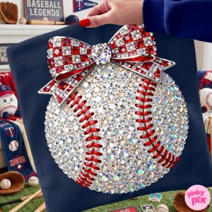 May include: A baseball-themed decorative pillow featuring a baseball design covered in sparkling crystals. A red and white crystal bow adorns the top. Red stitching details the baseball, set against a navy blue background. Baseball-themed decor is visible in the background.