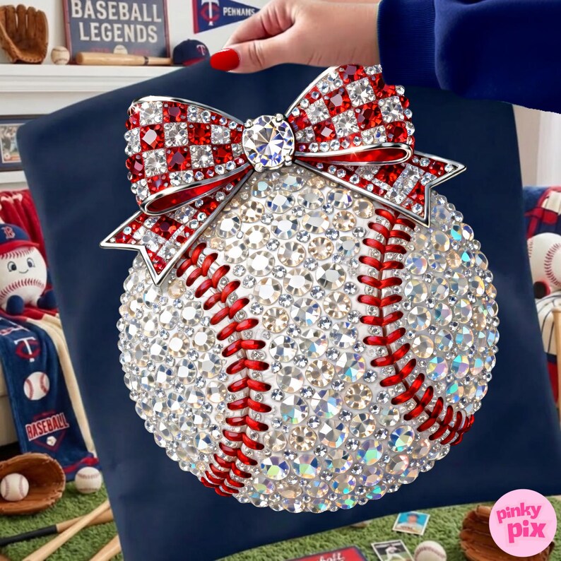 May include: A baseball-themed decorative pillow featuring a baseball design covered in sparkling crystals. A red and white crystal bow adorns the top. Red stitching details the baseball, set against a navy blue background. Baseball-themed decor is visible in the background.