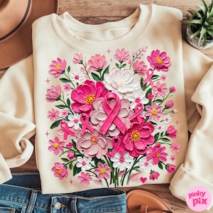 May include: A cream-coloured crewneck sweatshirt featuring a vibrant, textured floral print. The design includes various shades of pink and white flowers, green leaves, and multiple pink awareness ribbons integrated into the bouquet.